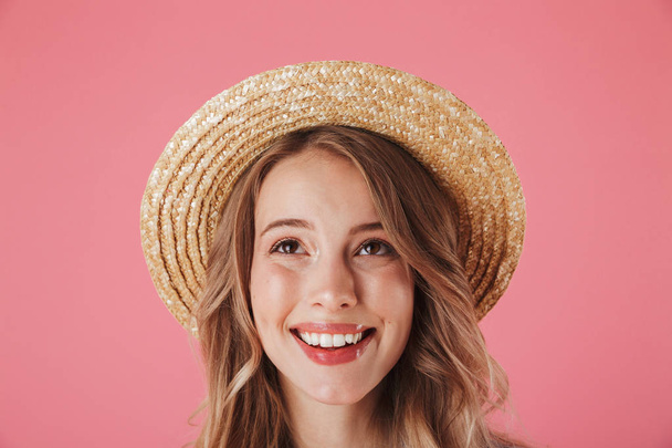 Close up portrait of a happy young woman in summer dress and straw hat looking up isolated over pink background - Foto, imagen