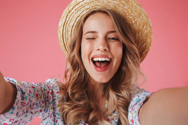 Close up portrait of a happy young woman in summer dress and straw hat taking a selfie isolated over pink background - Foto, immagini