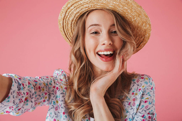 Close up portrait of a cheerful young woman in summer dress and straw hat taking a selfie isolated over pink background - Fotó, kép