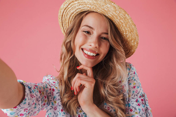Close up portrait of a beautiful young woman in summer dress and straw hat taking a selfie isolated over pink background - Foto, Imagem