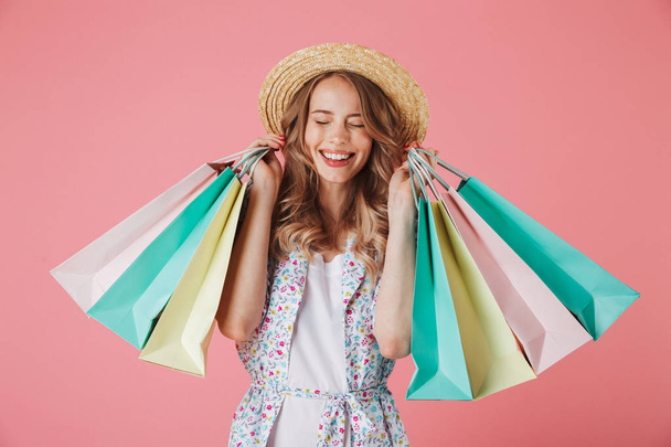 Portrait of a satisfied young woman in summer dress and straw hat holding shopping bags isolated over pink background - Foto, Imagem