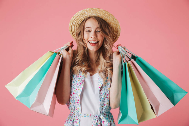 Portrait of a cheerful young woman in summer dress and straw hat holding shopping bags isolated over pink background - Foto, immagini