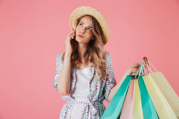 Portrait of a pensive young woman in summer dress and straw hat talking on mobile phone while holding shopping bags isolated over pink background - Foto, imagen