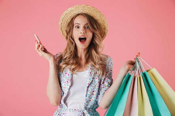 Portrait of a surprised young woman in summer dress and straw hat talking on mobile phone while holding shopping bags isolated over pink background - Fotoğraf, Görsel