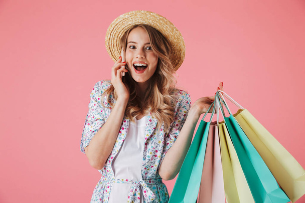 Portrait of a joyful young woman in summer dress and straw hat talking on mobile phone while holding shopping bags isolated over pink background - Photo, image