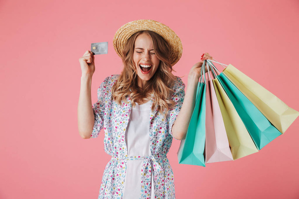 Portrait of an excited young woman in summer dress and straw hat showing plastic credit card while holding shopping bags isolated over pink background - Foto, imagen