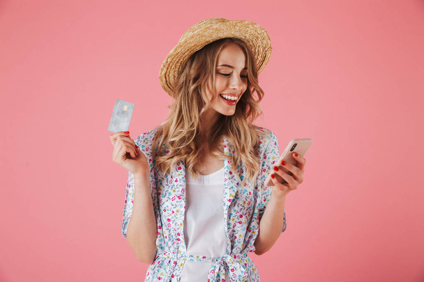 Portrait of a happy young woman in summer dress and straw hat showing plastic credit card while using mobile phone isolated over pink background - Φωτογραφία, εικόνα