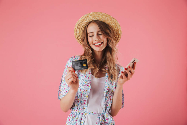 Portrait of a happy young woman in summer dress and straw hat looking at plastic credit card while using mobile phone isolated over pink background - Zdjęcie, obraz
