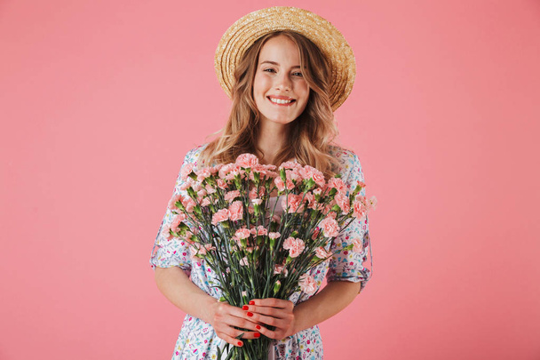 Portrait of a smiling young woman in summer dress and straw hat holding carnations bouquet isolated over pink background - Photo, Image