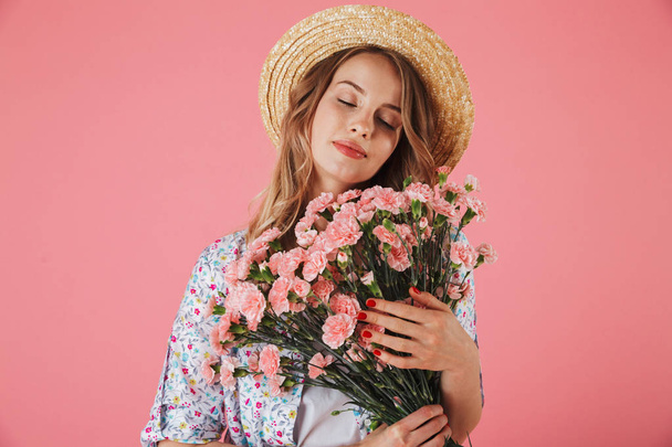 Portrait of a sensual young woman in summer dress and straw hat holding carnations bouquet isolated over pink background - Foto, Imagem