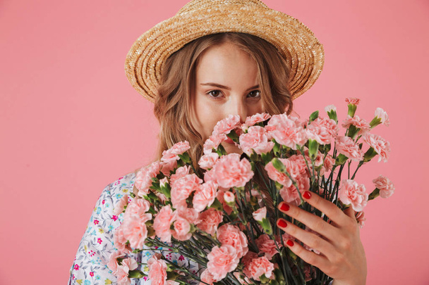 Close up portrait of a beautiful young woman in summer dress and straw hat smelling carnations bouquet isolated over pink background - Photo, Image