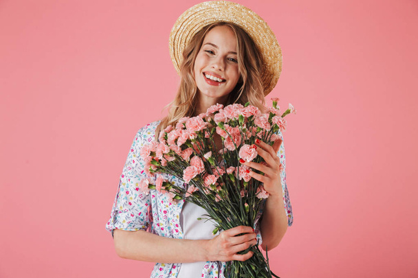 Portrait of a happy young woman in summer dress and straw hat holding carnations bouquet isolated over pink background - Foto, Imagen