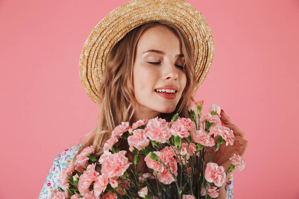 Close up portrait of a beautiful young woman in summer dress and straw hat holding carnations bouquet isolated over pink background - Foto, afbeelding