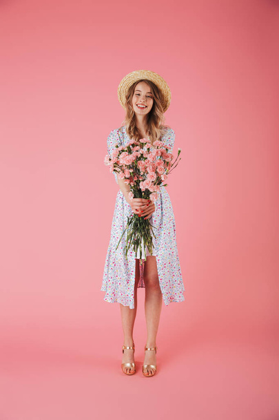 Full length portrait of a beautiful young woman in summer dress and straw hat posing while holding carnations bouquet isolated over pink background - Foto, imagen