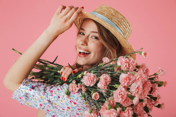 Close up portrait of a lovely young woman in summer dress and straw hat holding carnations bouquet isolated over pink background - Fotografie, Obrázek