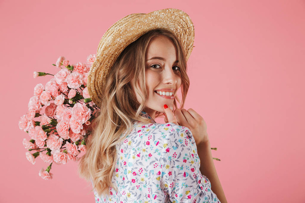 Close up portrait of a beautiful young woman in summer dress and straw hat holding carnations bouquet and looking over her shoulder isolated over pink background - Foto, Imagen