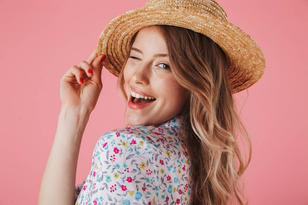 Close up portrait of a cheerful young woman in summer dress and straw hat posing and looking at camera isolated over pink background - Фото, зображення