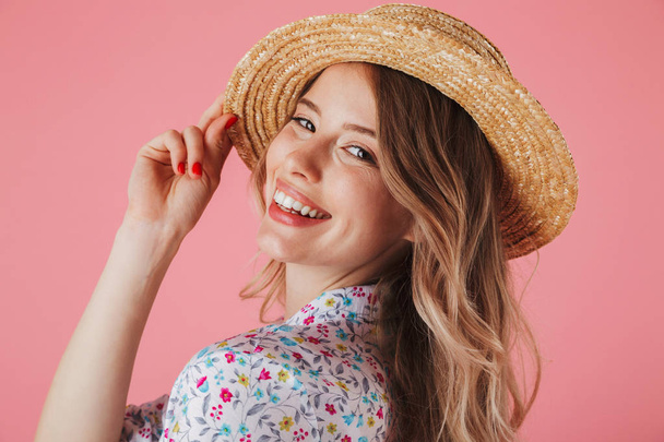 Close up portrait of a pretty young woman in summer dress and straw hat posing and looking at camera isolated over pink background - Foto, Bild