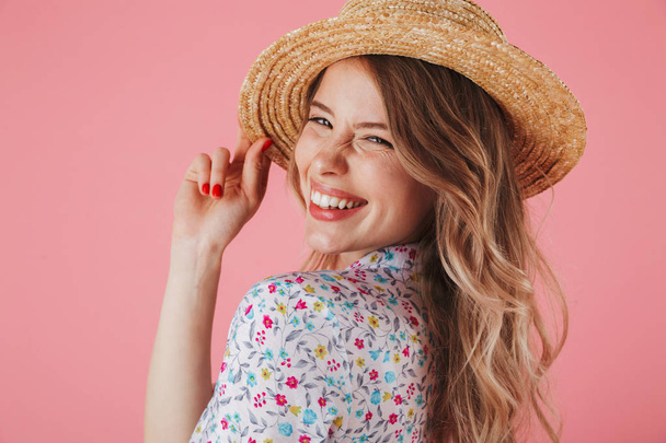Close up portrait of a laughing young woman in summer dress and straw hat posing and looking at camera isolated over pink background - Valokuva, kuva