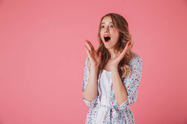 Portrait of an amazed young woman in summer dress looking away at copy space isolated over pink background - Foto, afbeelding
