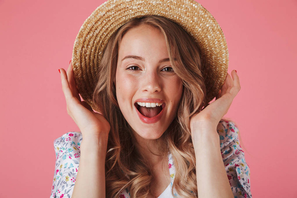 Close up portrait of a happy young woman in summer dress and straw hat looking at camera isolated over pink background - Zdjęcie, obraz