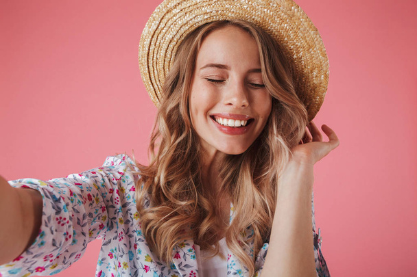 Close up portrait of a lovely young woman in summer dress and straw hat taking a selfie isolated over pink background - Photo, image