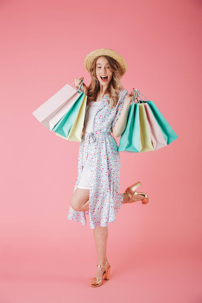 Full length portrait of a cheerful young woman in summer dress and straw hat holding shopping bags and celebrating isolated over pink background - Foto, Imagen
