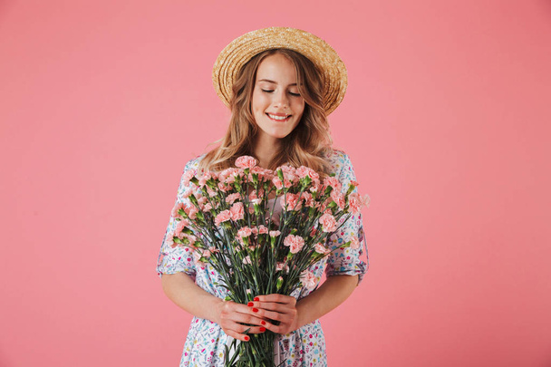 Portrait of a lovely young woman in summer dress and straw hat holding carnations bouquet isolated over pink background - Fotoğraf, Görsel
