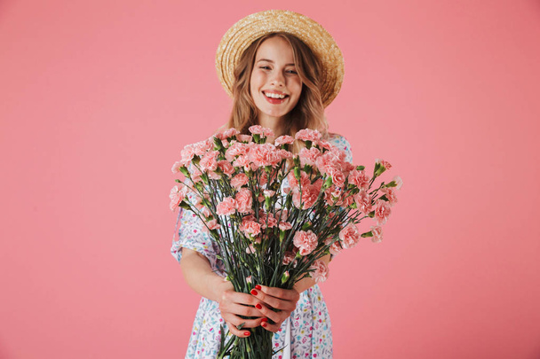 Portrait of a cheerful young woman in summer dress and straw hat giving carnations bouquet isolated over pink background - Photo, Image