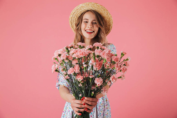 Portrait of a happy young woman in summer dress and straw hat giving carnations bouquet isolated over pink background - Фото, зображення