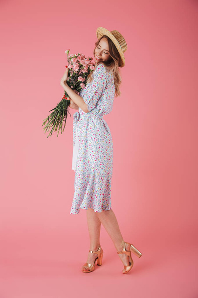 Full length portrait of a lovely young woman in summer dress and straw hat posing while holding carnations bouquet isolated over pink background - Φωτογραφία, εικόνα