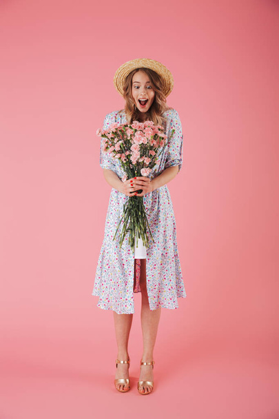 Full length portrait of a cheerful young woman in summer dress and straw hat posing while holding carnations bouquet isolated over pink background - Φωτογραφία, εικόνα