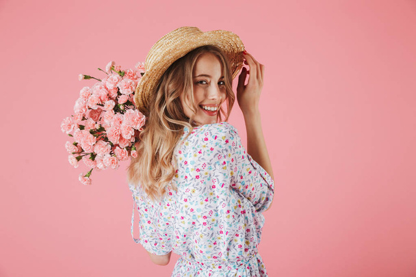 Close up portrait of an attractive young woman in summer dress and straw hat holding carnations bouquet and looking over her shoulder isolated over pink background - Photo, image