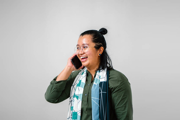 Malay woman wearing olive green shirt, spectacles and scarf smiling talking on smartphone - Photo, image