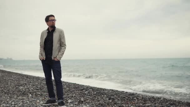 człowiek w okularach stoi na kamienistych plaży, w pobliżu morza i wygląda odległości - Materiał filmowy, wideo