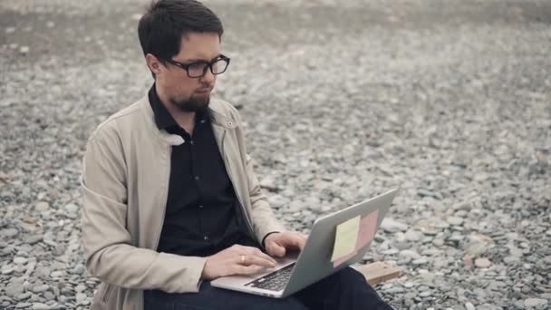 νεαρός επιχειρηματίας αναπτύσσει ένα εκκίνησης σε μια βραχώδη παραλία, χρησιμοποιώντας ένα φορητό υπολογιστή - Πλάνα, βίντεο