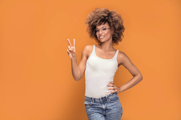 Photo of beautiful, smiling, positive afro girl showing peace symbol, looking at camera, posing on colorful background. - Φωτογραφία, εικόνα