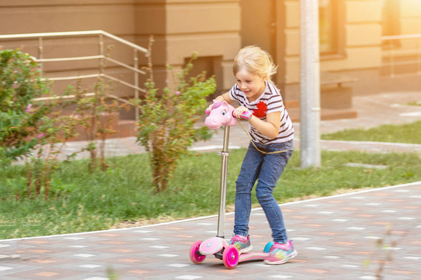 Cute little girl riding scooter in city park on bright summer day. Blond toddler having fun on street. Active leisure and outdoor sport for children concept. - Foto, afbeelding
