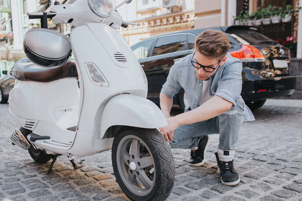 A picture of guy sitting in squat position and looking at motorcycles wheel. He is trying to fix it. Guy looks worried and serious. - Foto, Bild