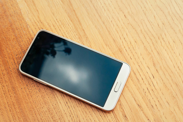 Vista superior de un solo teléfono inteligente blanco con pantalla en blanco negro para su diseño en mesa de madera clara. Cielo y flores reflejadas en la pantalla
 - Foto, Imagen