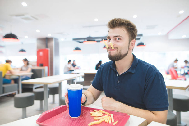 Vicces ember ül egy gyorsétterem egy pohár ital a kezében és a krumplit a szájában, és úgy néz ki, a kamera. Szakáll vicces ember eszik gyorsételek. Fast Food koncepció. - Fotó, kép