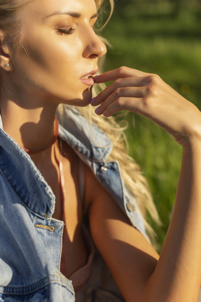 Beautiful blonde slim girl wearing pink lingerie and jeans jacket sits on the grass in the park outdoors. Summer sunny lifestyle photo. Advertising and conceptual design. Copy space. - Φωτογραφία, εικόνα