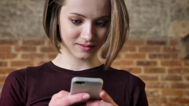 Young beautiful girl is reading message on her smartphone, she is surprised, communication concept, brick background - Imágenes, Vídeo