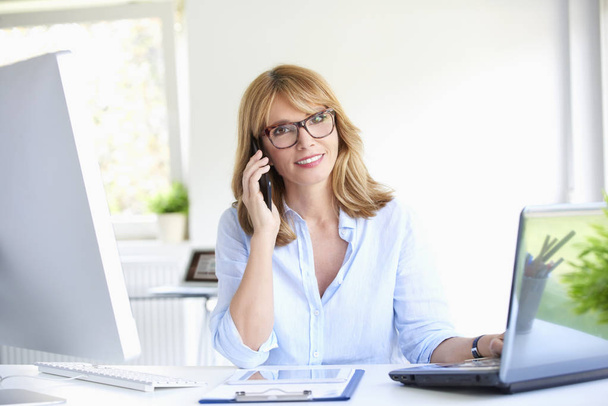 Close-up portrait of attractive mature businesswoman talking with her client on mobile phone while sitting at office desk working on laptop. - Foto, Imagem