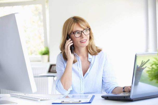 Close-up portrait of attractive mature businesswoman talking with her client on mobile phone while sitting at office desk working on laptop. - Foto, Bild