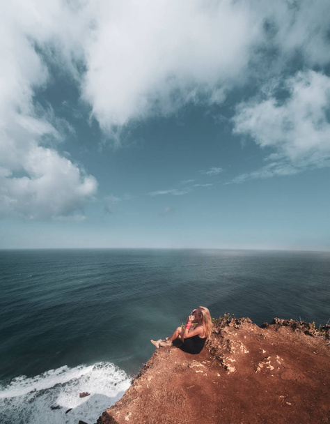 Pretty girl sitting on the cliff with amazing ocean and cloudy sky view  - Fotó, kép