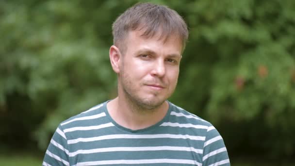 Jonge knappe man dragen gestreept t-shirt - emotionele portret buitenshuis in het park op groene achtergrond - close-up. - Video