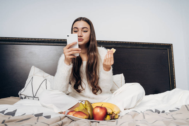 cute long-haired girl in white pajamas sitting in bed early in the morning, eating fruits and looking into her smartphone - Photo, image