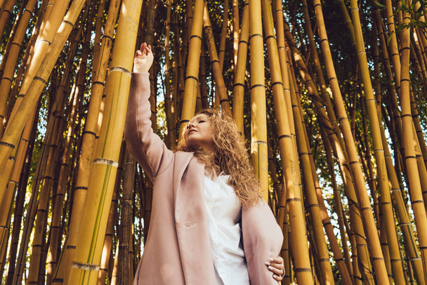 stylish curly woman in a pink coat enjoying the nature in the park, next to the tall bamboo trees - Foto, afbeelding