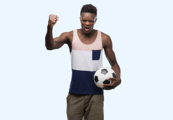 Jonge african american man die voetbal voetbal geërgerd en gefrustreerd schreeuwen met woede, gek en schreeuwen met opgeheven hand, woede concept - Foto, afbeelding